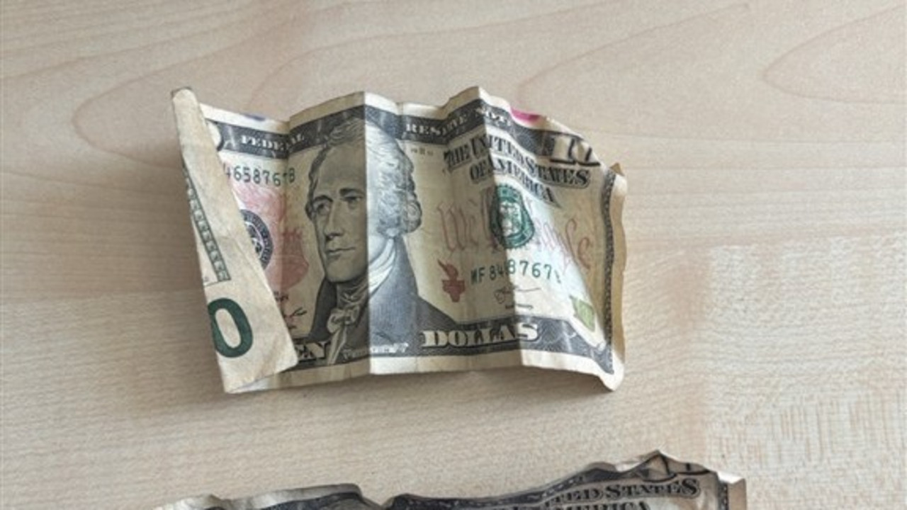Das Foto zeigt zwei 10-Dollarnoten. Ein Schein wird von einer Hand mit blauem Handschuh auf einen Tisch angedrückt.