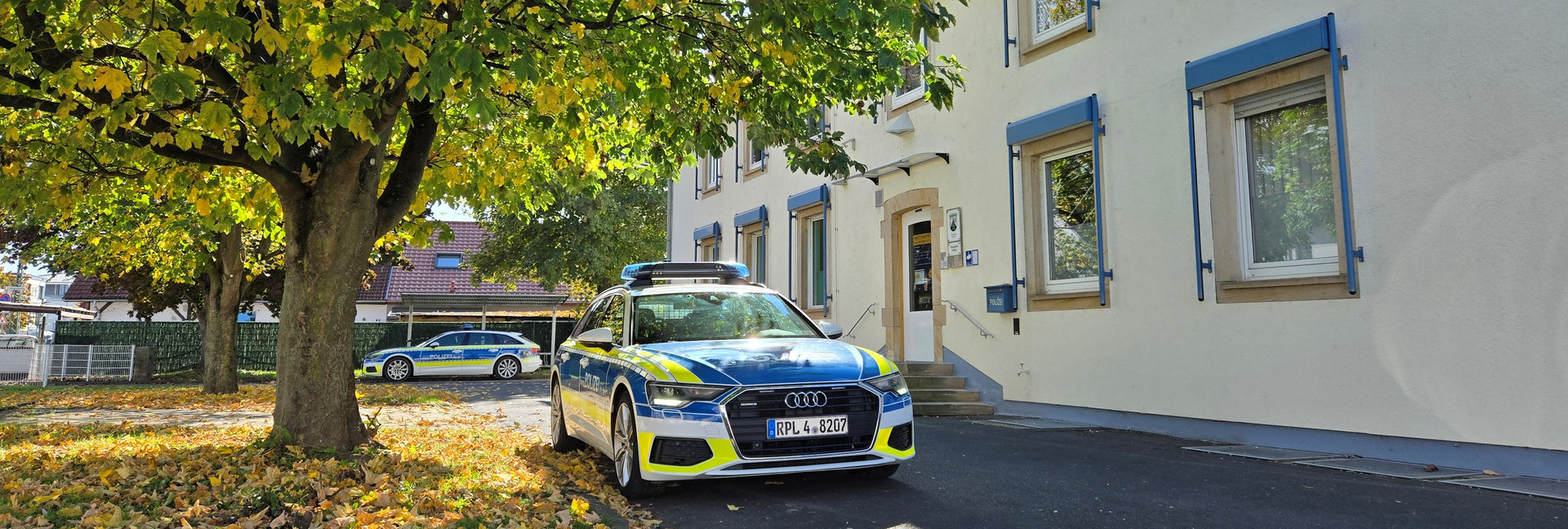 Zwei Polizeiautos stehen vor einem hellen Gebäude mit blauen Fensterläden, vermutlich einer Polizeistation. Ein Audi-Polizeiwagen parkt nahe dem Eingang, ein weiteres Fahrzeug etwas weiter hinten. Vor dem Gebäude wächst ein großer Baum mit gelb-grünem Herbstlaub, am Boden liegen viele Blätter.