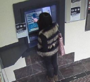 Eine Frau steht vor dem Geldautomaten. Sie trägt eine gestreifte Jacke und hat eine Umhängetasche über der Schulter hängen.