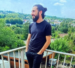 Foto eines Mannes mit dunklem Bart, dunklen, zu einem Pferdeschwanz gebundenen Haaren, schwarzem T-Shirt, schwarzer,kurzer Hose und schwarzen Turnschuhen.