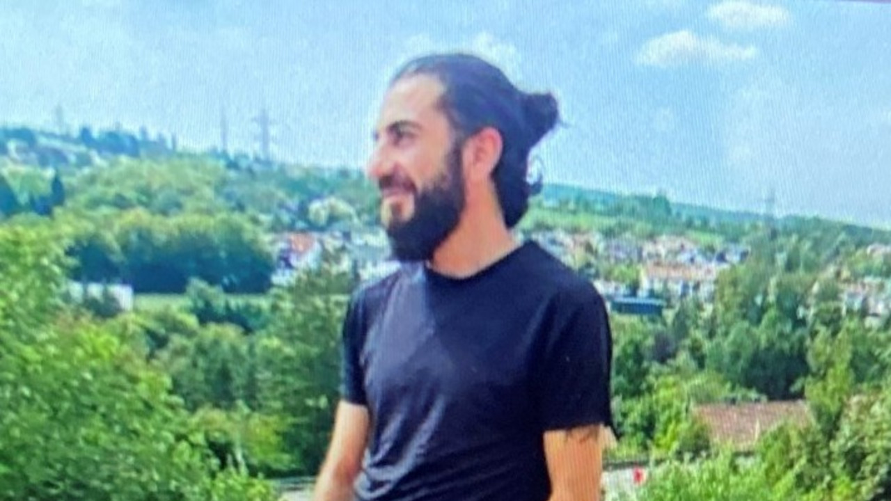 Foto eines Mannes mit dunklem Bart, dunklen, zu einem Pferdeschwanz gebundenen Haaren, schwarzem T-Shirt, schwarzer,kurzer Hose und schwarzen Turnschuhen.