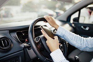 Eine Frau hält mit der rechten  Hand das Lenkrad, mit der linken Hand tippt sie etwas in ihrem Smartphone