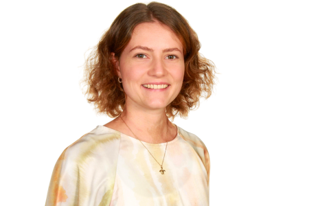 Eine Person mit schulterlangem, lockigem braunem Haar lächelt freundlich in die Kamera. Sie trägt ein helles Oberteil mit dezentem Muster, eine Halskette mit einem kleinen Kreuzanhänger und große, runde Ohrringe. Der Hintergrund ist einfarbig weiß, wodurch die Person deutlich hervorgehoben wird.